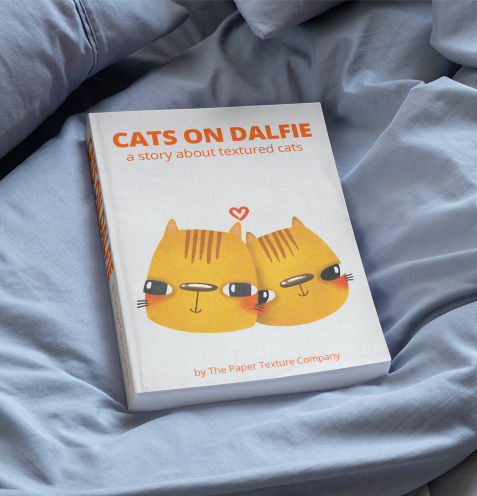 A book titled "Cats on Dalfie: a story about textured cats" with an illustration of two cartoon cats and a small red heart is lying on light blue bed sheets. The author is The Paper Texture Company.