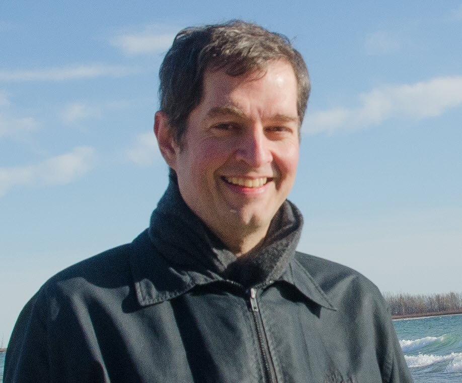 Digital paper texture - A man with short dark hair wearing a black jacket and scarf is standing outdoors on a sunny day. The sky is blue with some clouds, and there is water and a tree line in the background.